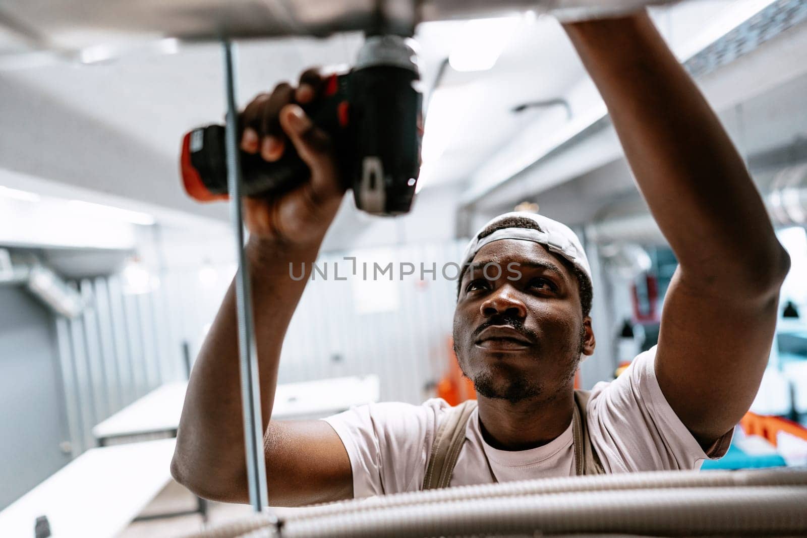 A construction worker uses a power drill to secure fixtures in a commercial kitchen. Bright daylight shines in as workers tackle renovation tasks in a bustling environment.