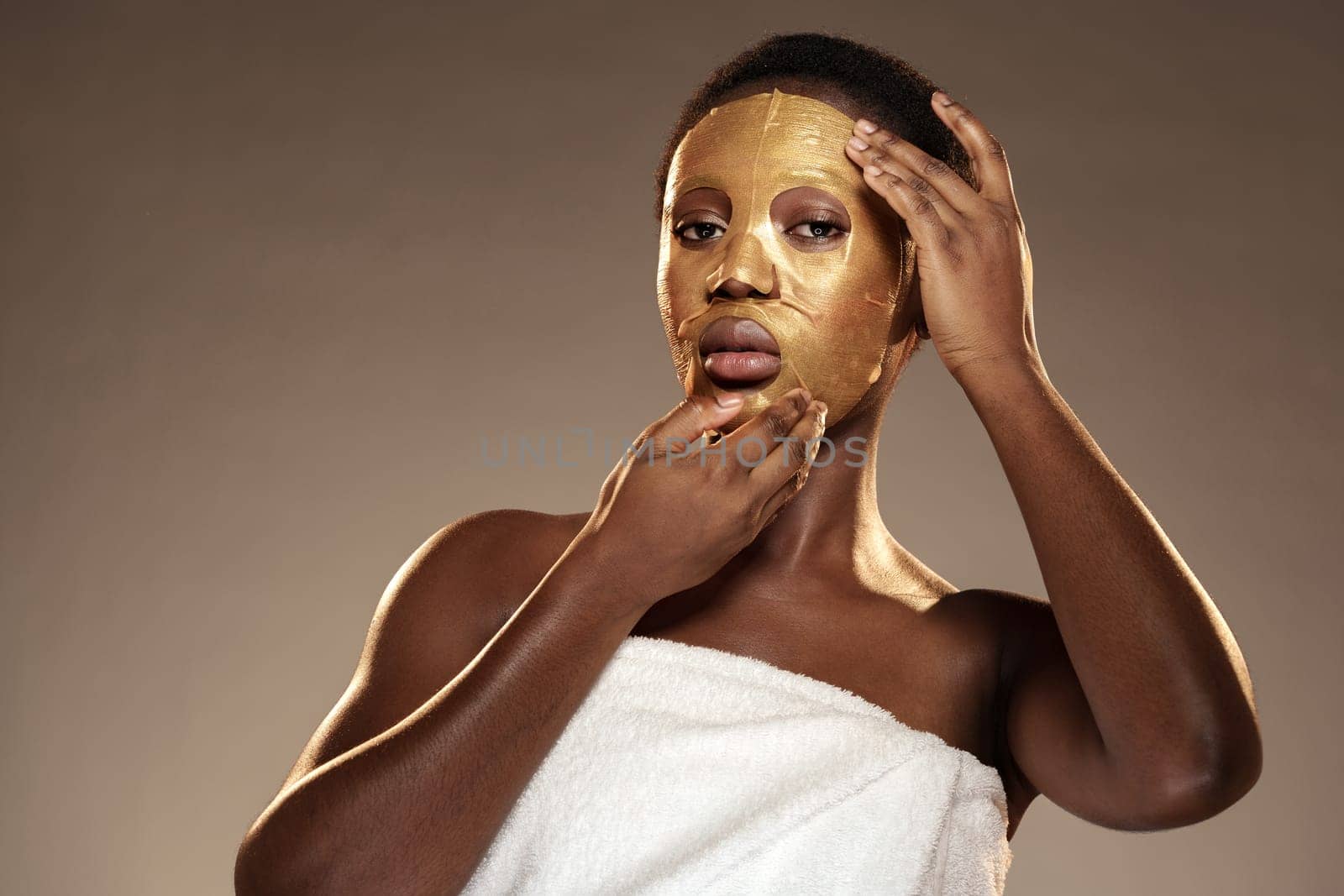 A woman in a towel gently removes a golden facial mask from her face, showcasing her radiant skin. The calm indoor atmosphere emphasizes relaxation and wellness.