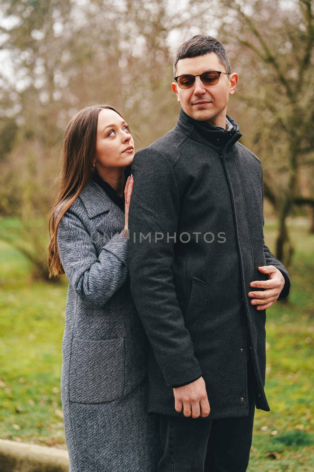 A woman in a gray coat affectionately leans against a man dressed in a black jacket, both smiling while surrounded by green trees in a park setting.
