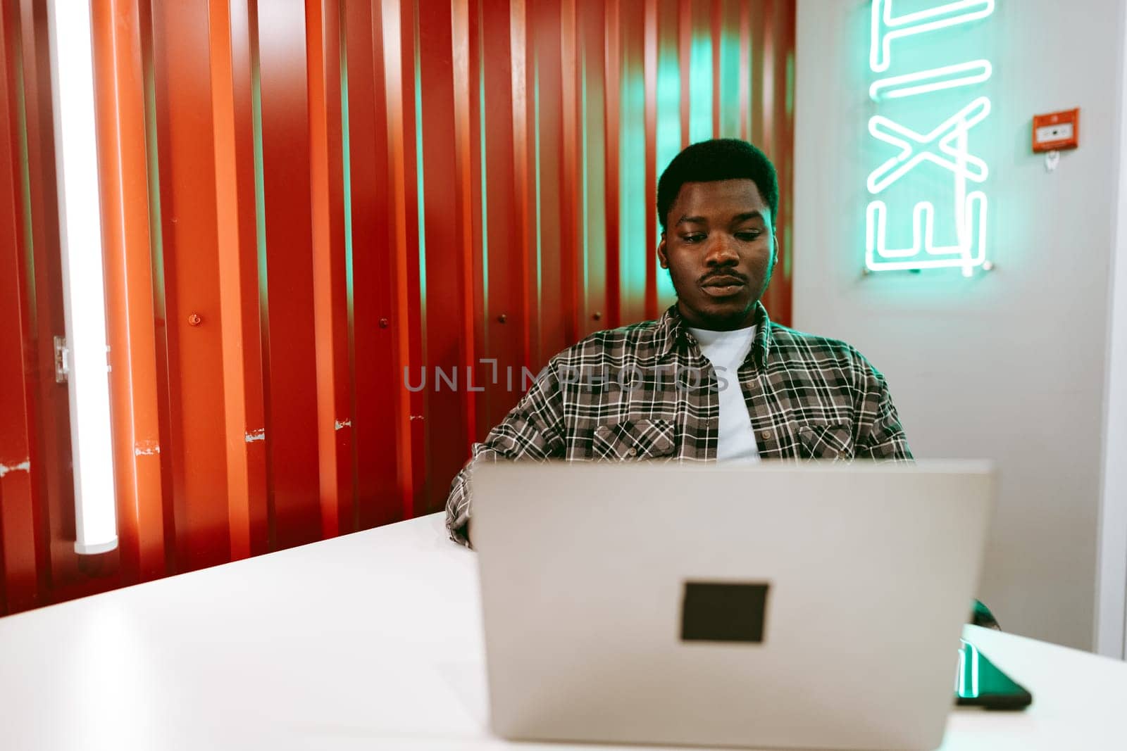 A focused young man sits at a minimalistic table, using his laptop in a lively coworking space with bright orange walls and glowing green exit signage.
