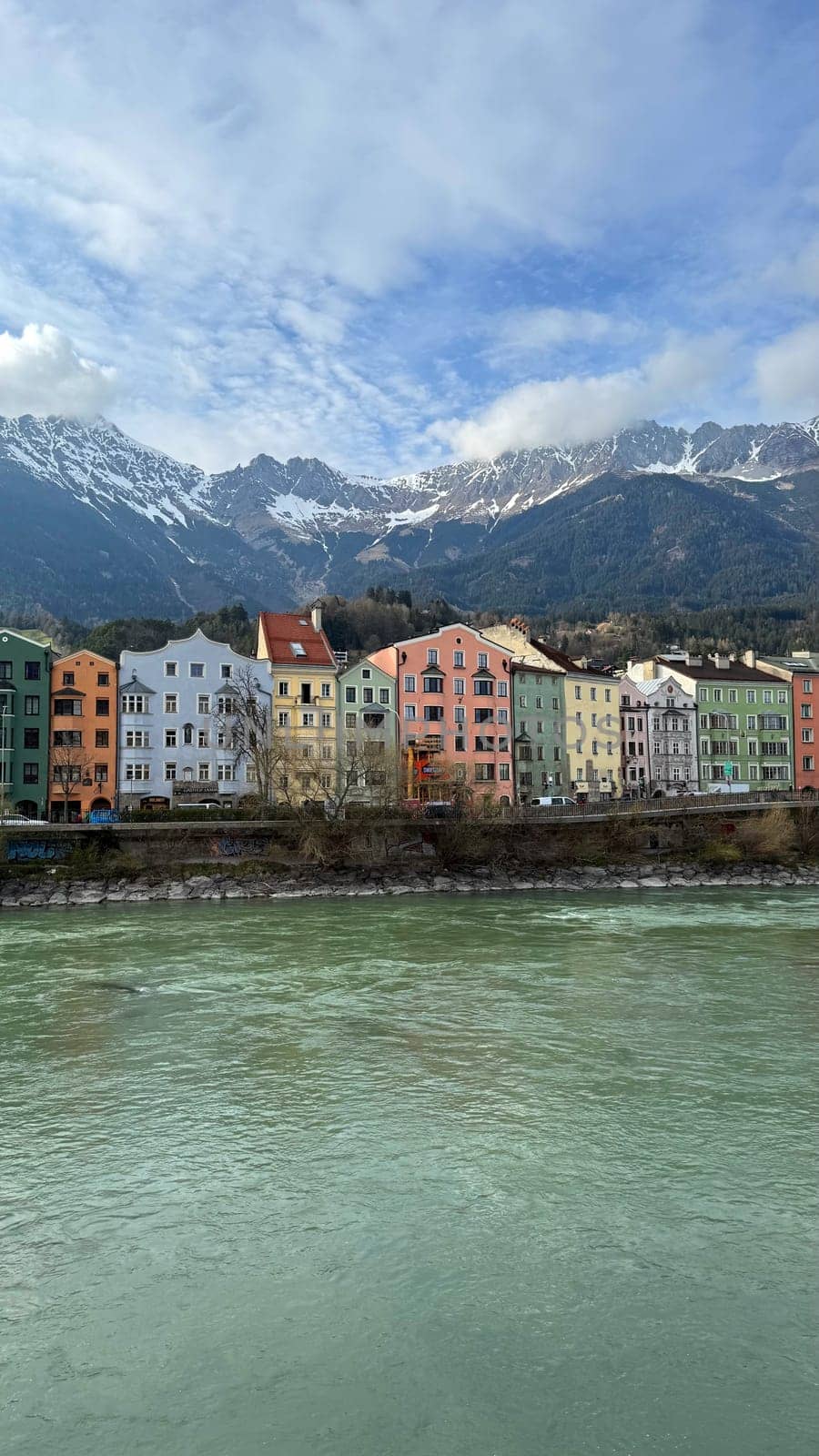 Innsbruck, Austria - March 28, 2025: Charming pastel-colored buildings line a riverbank Inn against a backdrop of snow-capped Alpine mountains and clear blue sky, providing a picturesque view of a serene and vibrant European town.