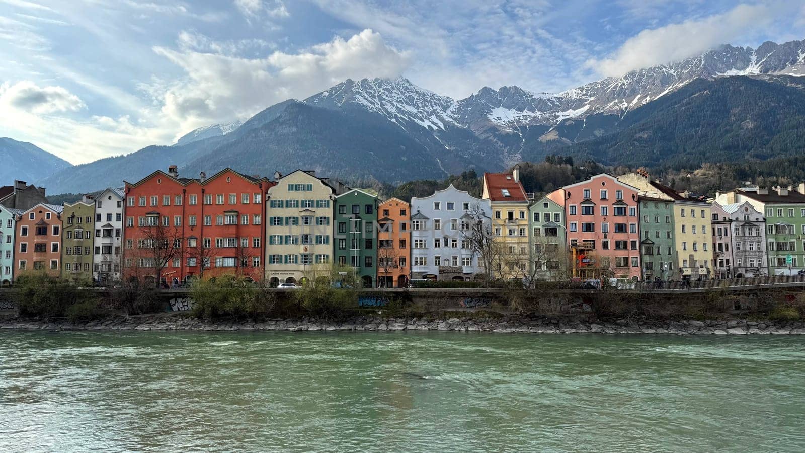 Innsbruck, Austria - March 28, 2025: Vibrant row of colorful houses along the Inn River in Innsbruck, Austria, with snow-capped mountains in the background. A picturesque European cityscape in spring light, blending traditional architecture and natural alpine beauty.