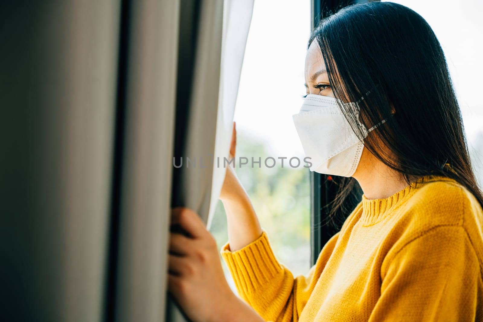 A young Asian woman in a medical mask looks through the window feeling upset and distressed due to COVID-19. Reflecting fear isolation and pandemic-related anxiety.
