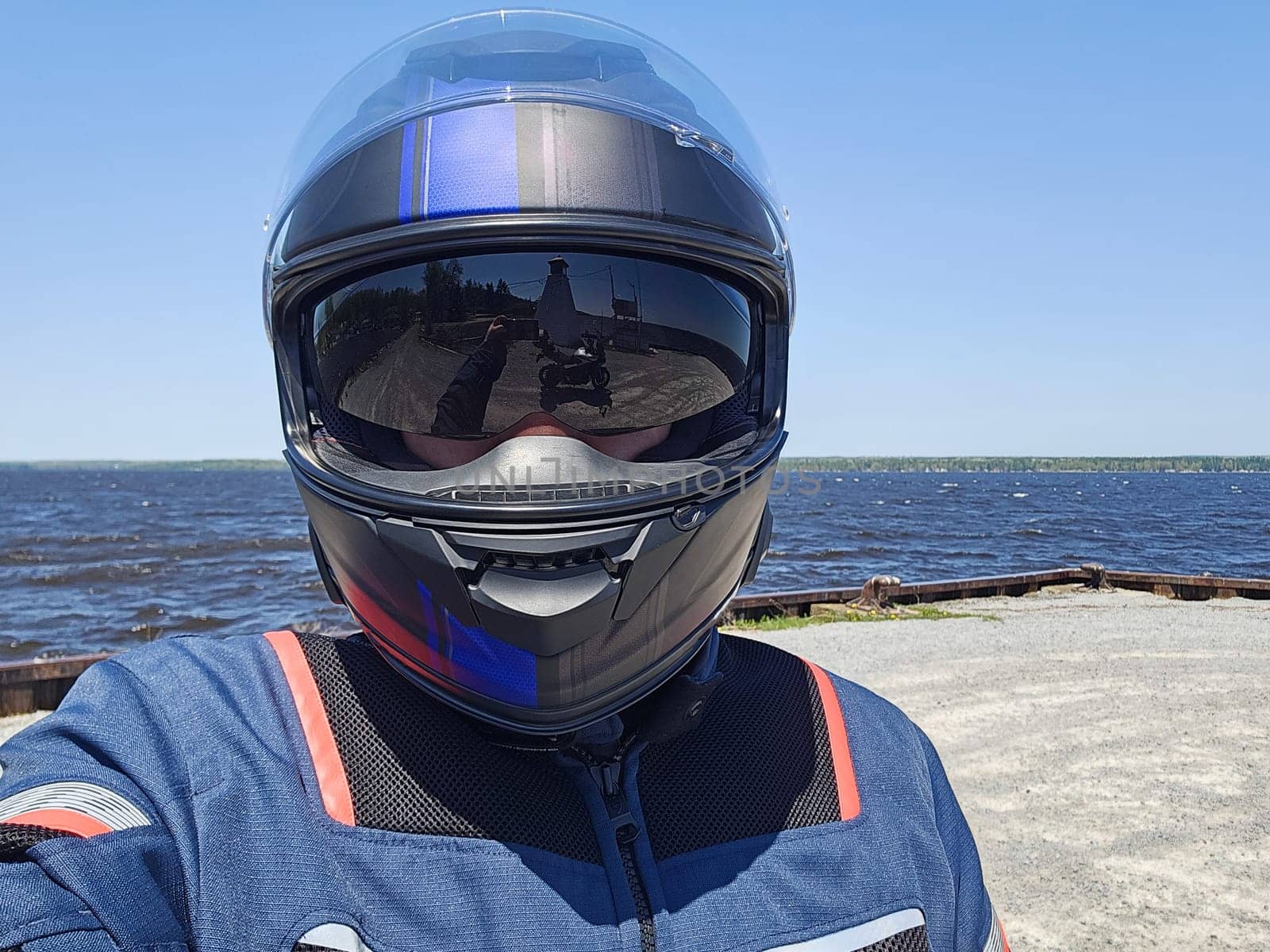 A motorcycle rider takes a selfie wearing a dark helmet with a reflective visor. The scenic lake and sunny day create a beautiful backdrop for the rider in a blue and black jacket.