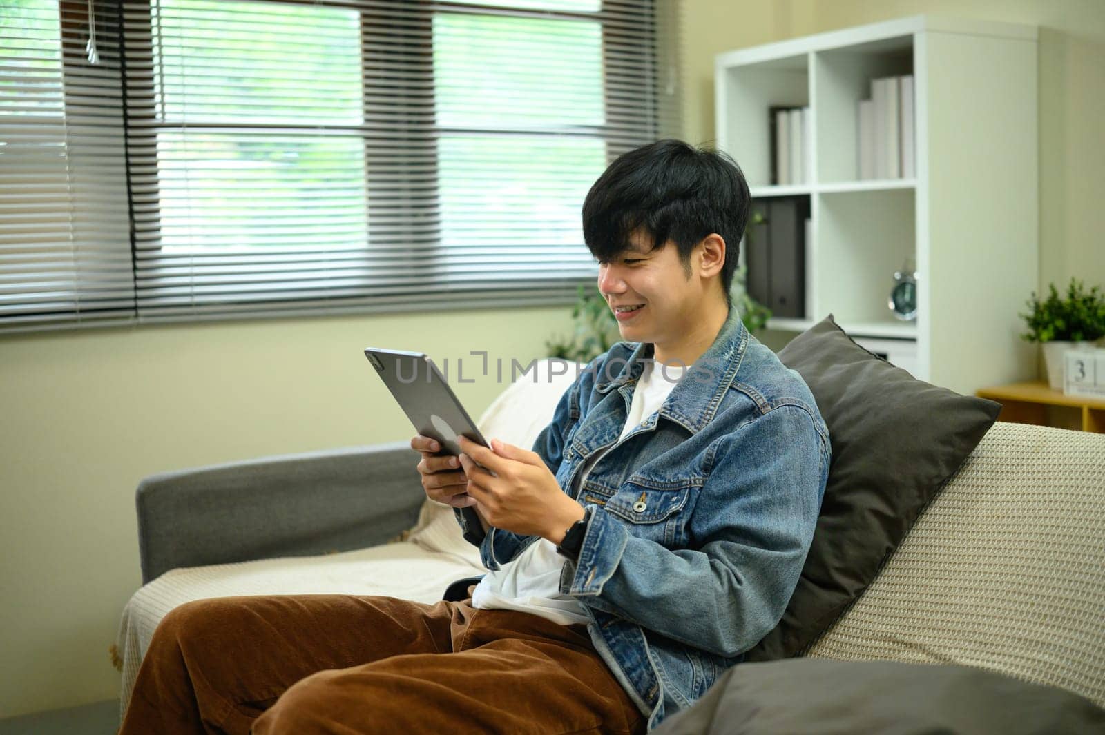 Happy young asian man wearing denim jacket using digital tablet in living room.
