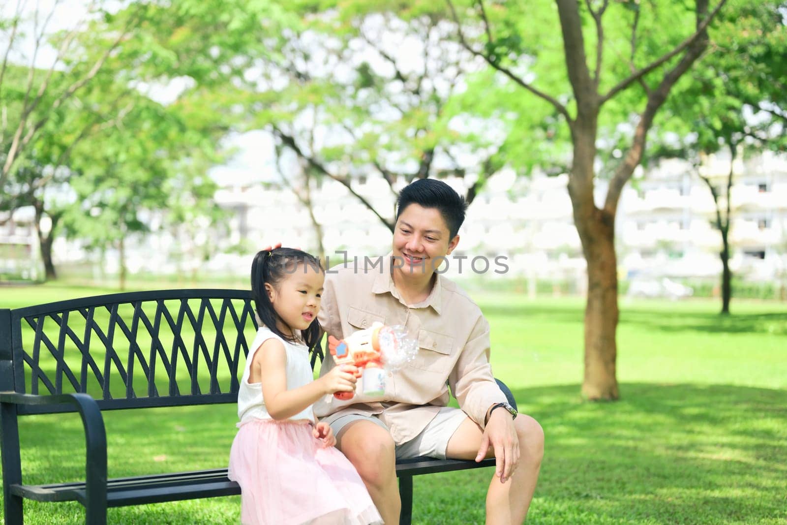 Father and daughter sit on a park bench enjoying a bubble toy on a sunny day.