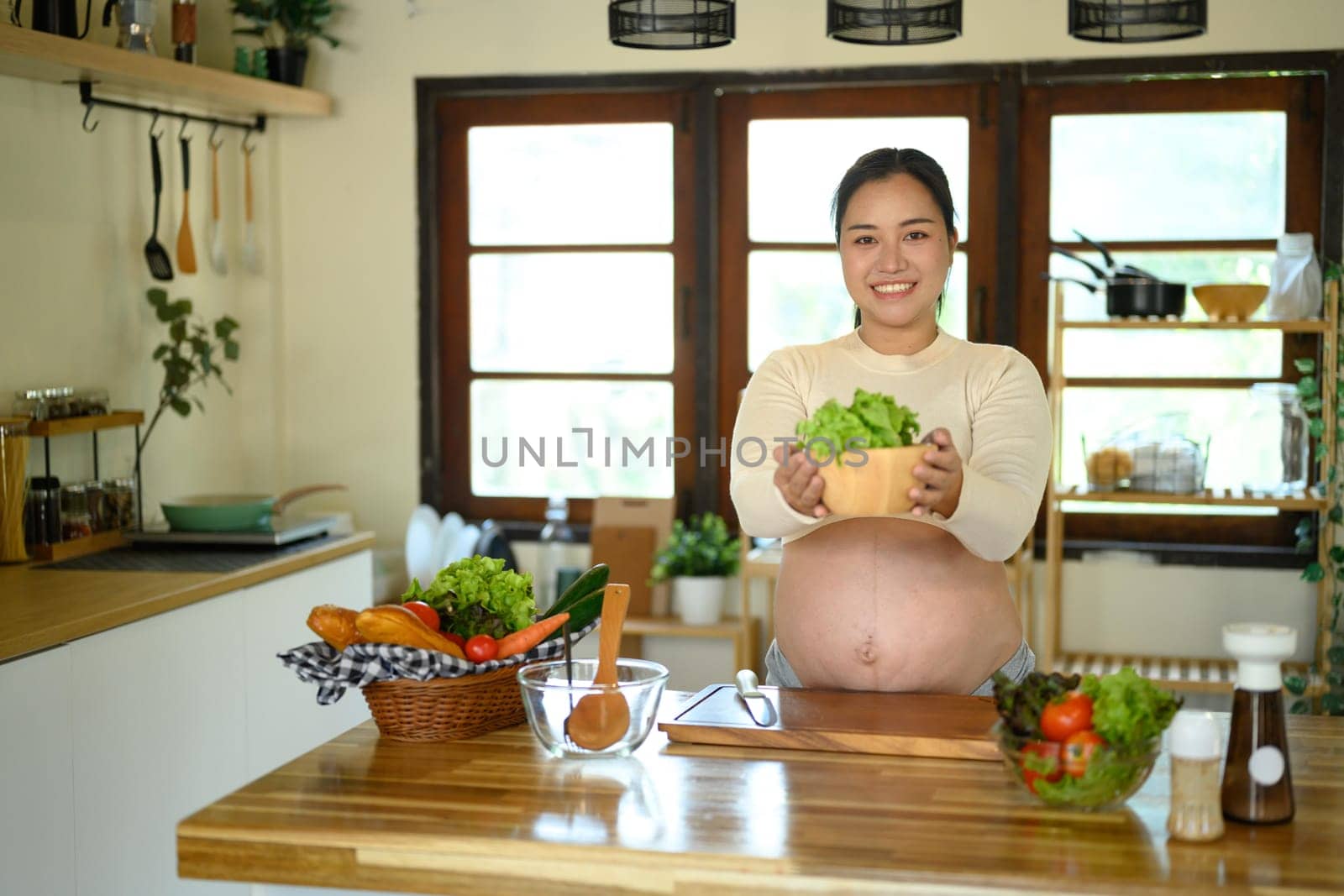 Asian pregnant woman standing in a bright kitchen, presenting a bowl of salad.