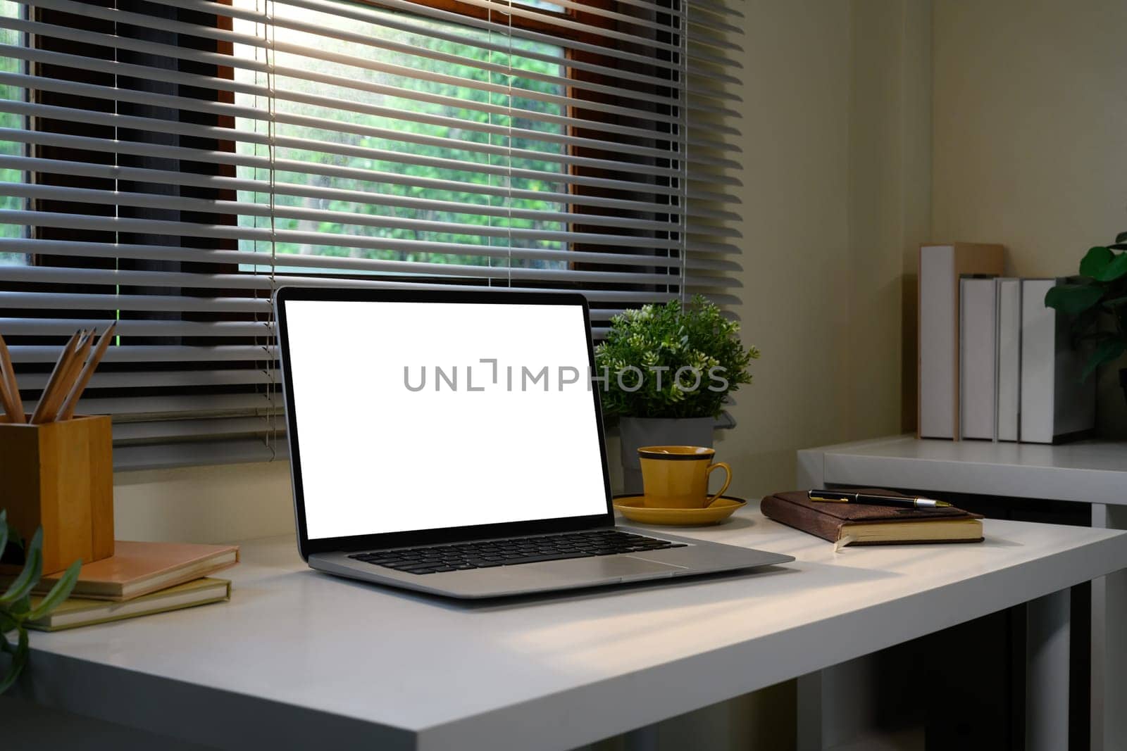 Modern home office desk with a blank laptop screen, notebook, and plants by the window.