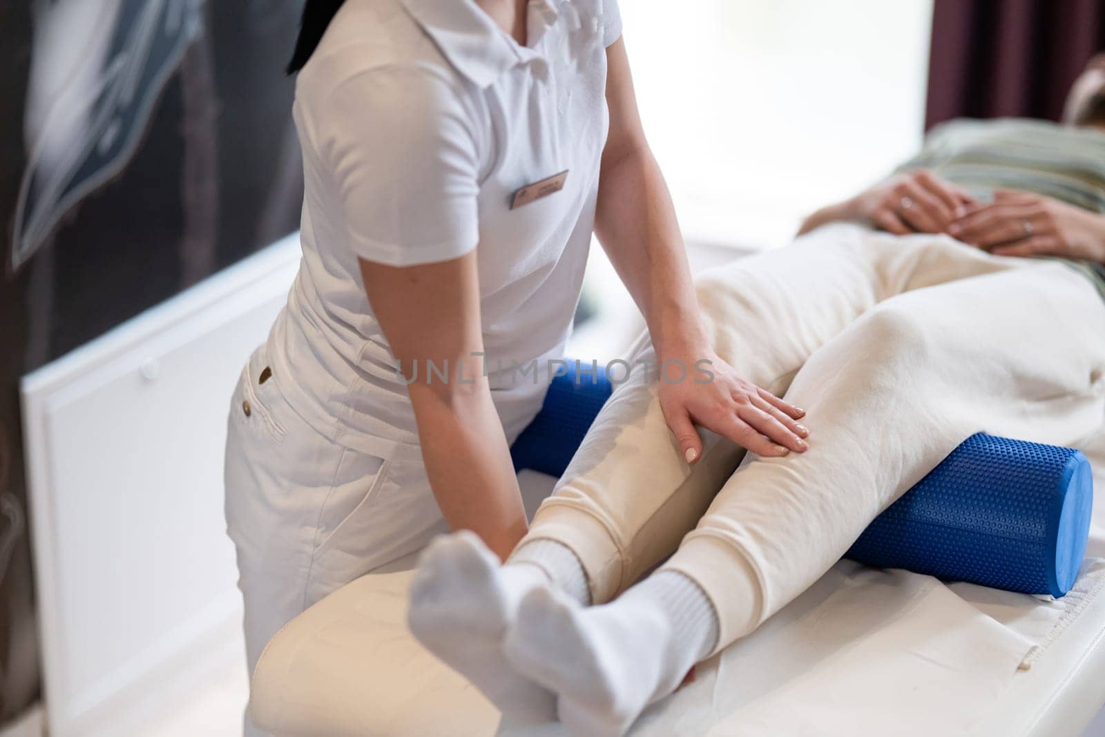 A therapist guiding a patient through leg exercises with the help of a blue foam roller in a therapy clinic.