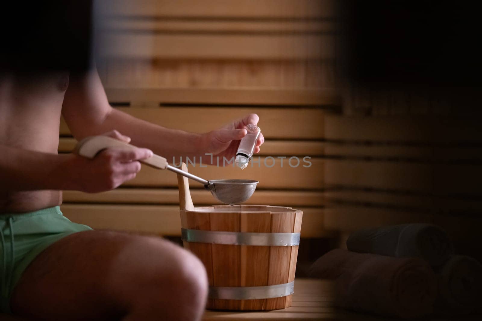Close up of a mans hand holding essential oils inside a traditional wooden sauna with bucket and ladle.