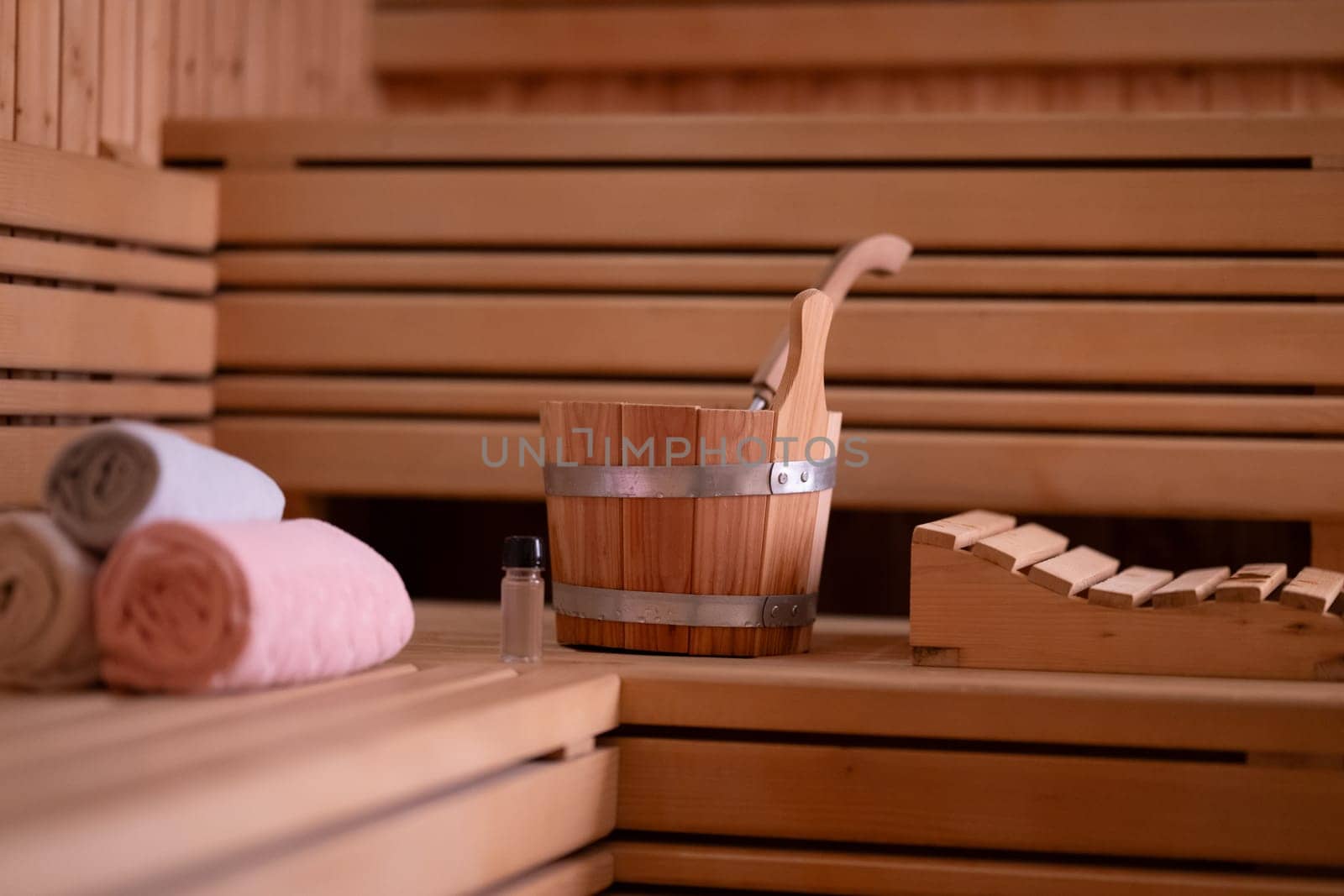 Detailed close up of sauna bucket with ladle and essential oils next to rolled towels on wooden bench inside sauna.