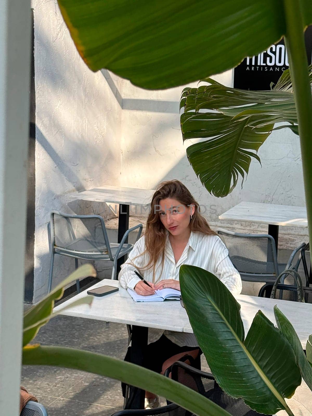 A woman sits at a table in an outdoor cafe, writing in a notebook, with tropical plants around her. The sun shines softly.