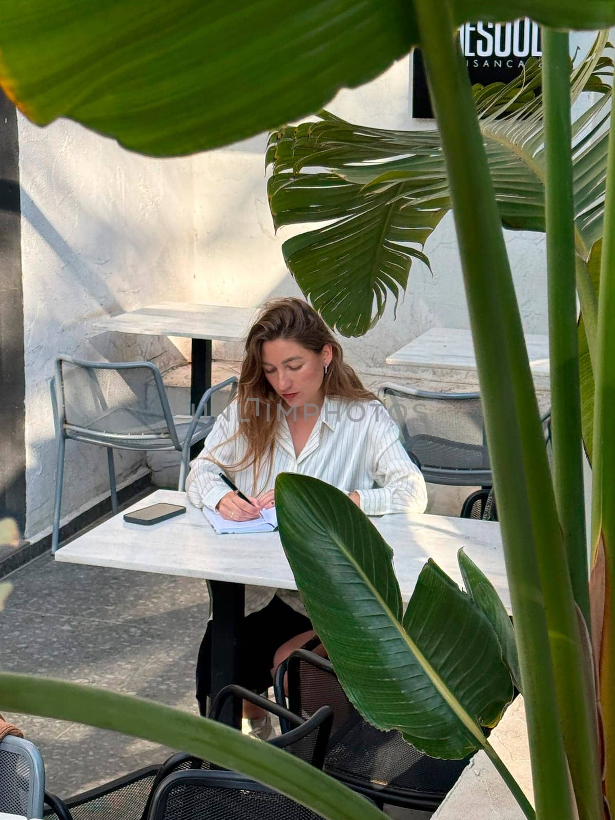 A young woman focuses on her studies at an outdoor cafe, surrounded by plants and enjoying a sunny afternoon.