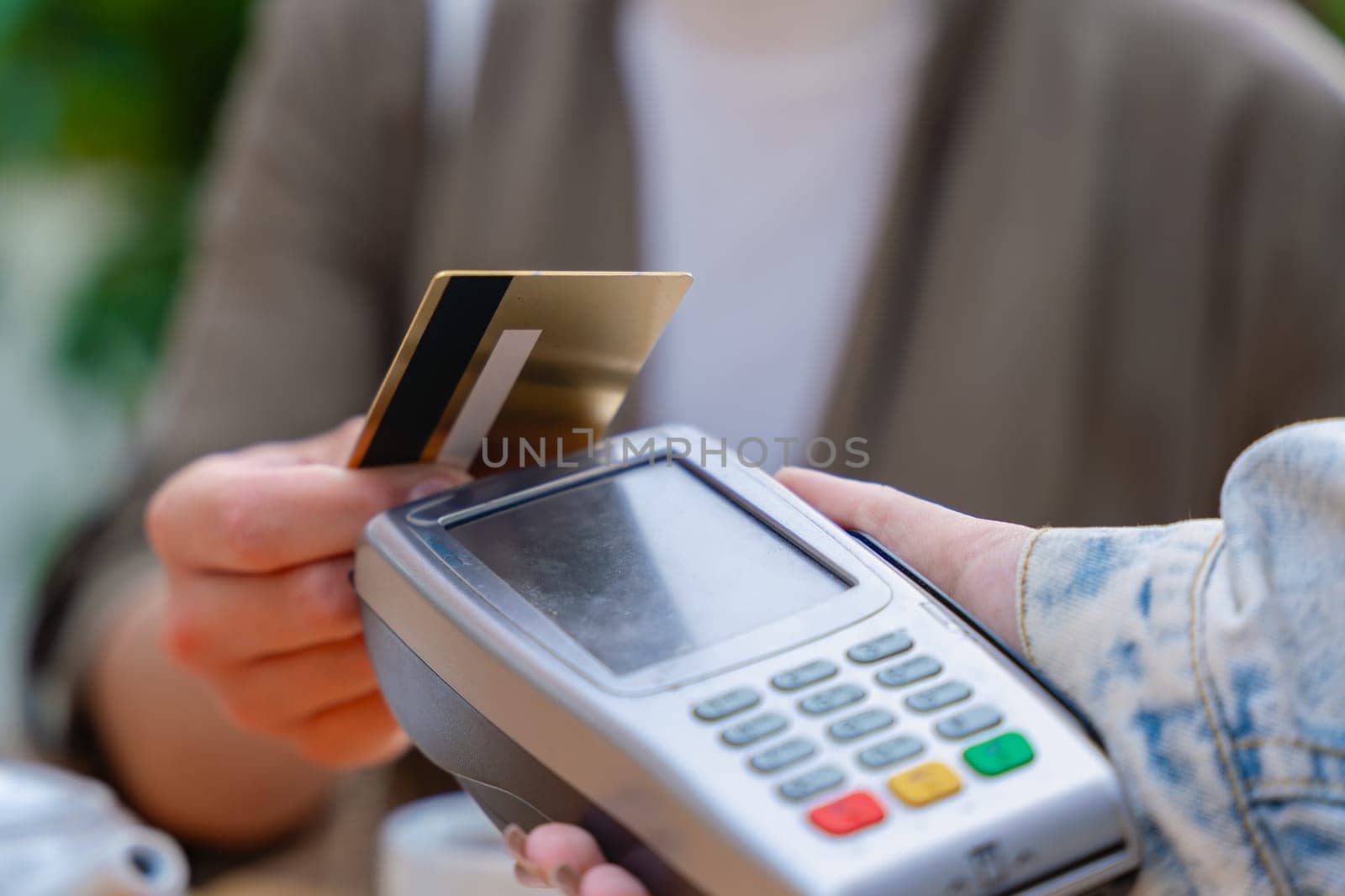 Close-up view of a person making a contactless payment using a credit card on a POS terminal. The scene takes place in a cafe setting with a teapot in the background, showing modern transaction ease