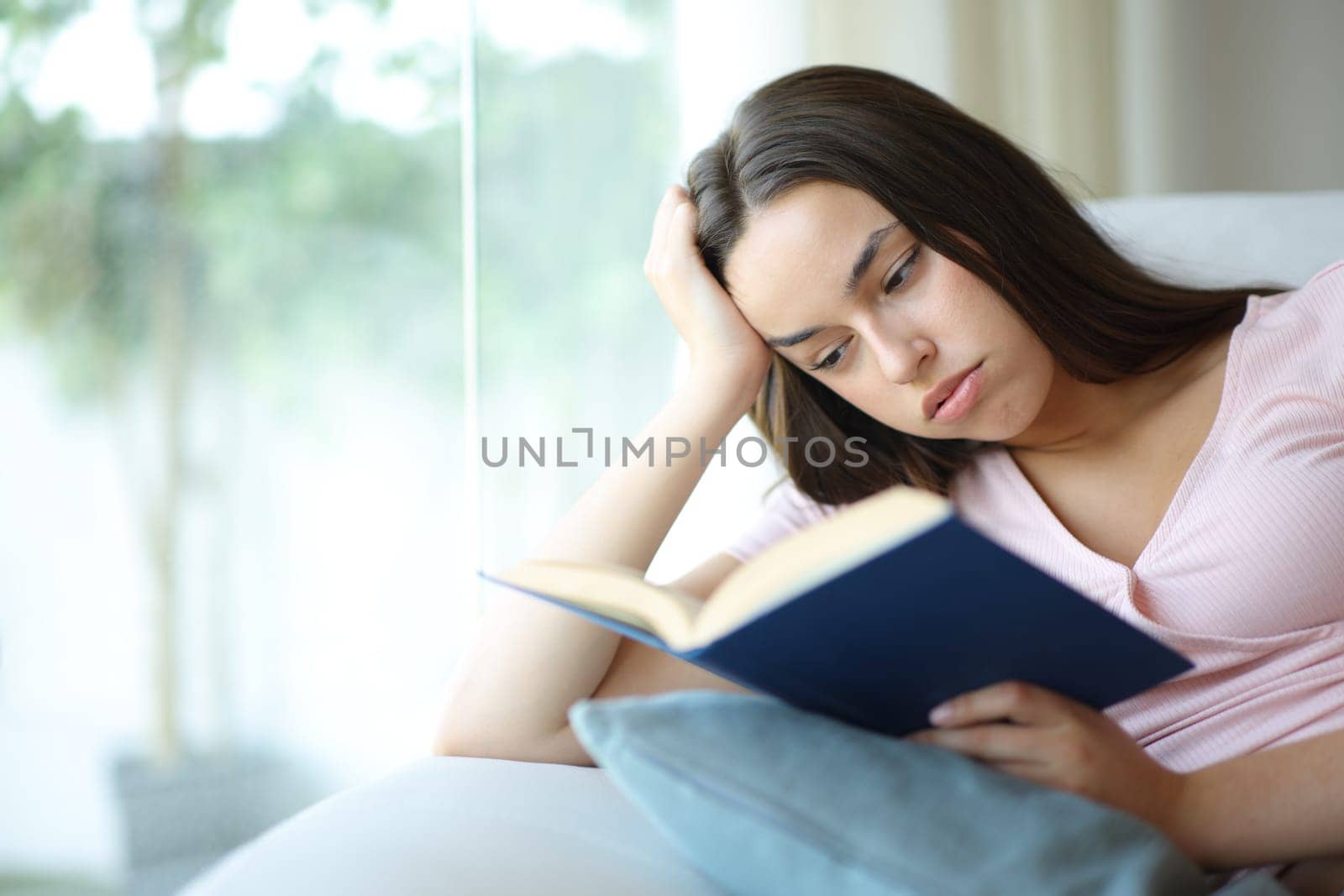 Bored woman is reading a paper book sitting on a couch at home