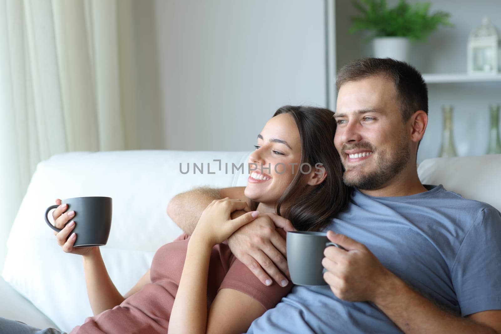Happy couple drinking tea looking away sitting on a couch in a house interior