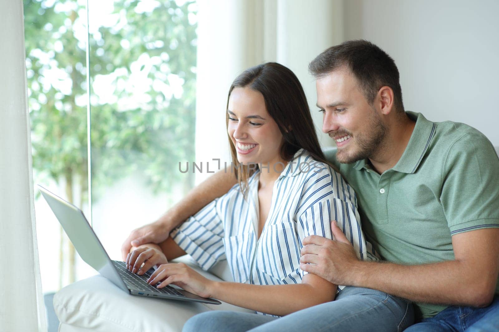 Happy couple using a laptop together sitting in a couch in a house interior
