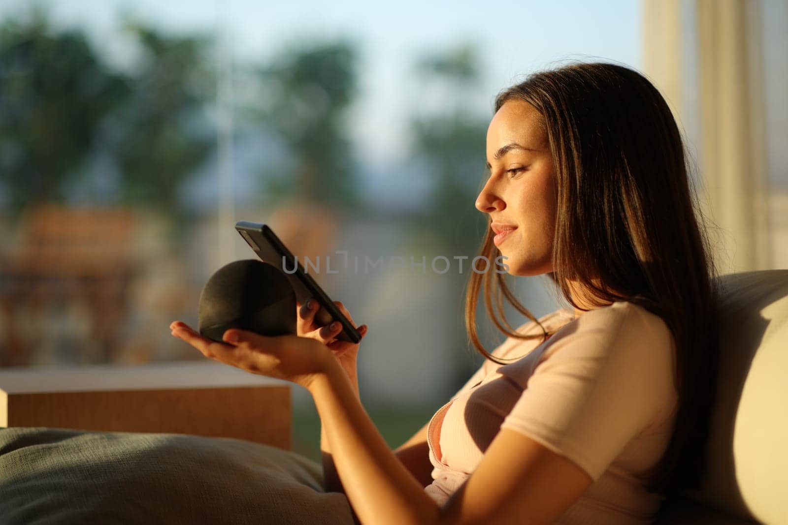 Woman pairing phone with smart speaker at sunset sitting on a couch at home