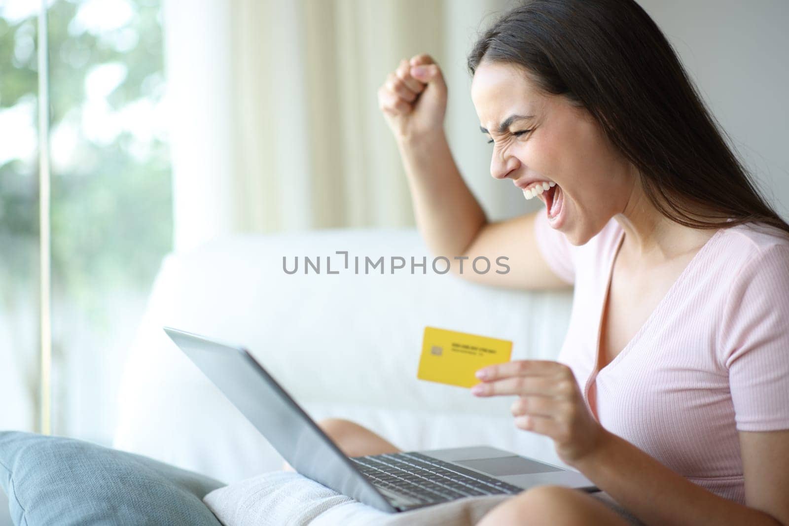 Woman celebrating exciting discount buying online with laptop and credit card sitting on a couch at home