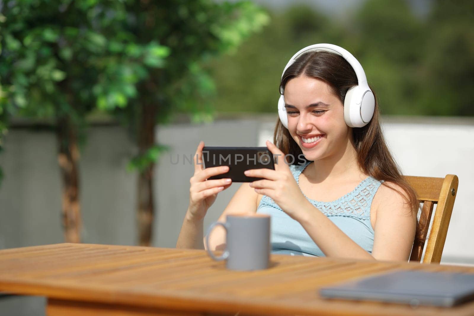 Happy woman wearing headphone in a terrace watching online media on phone a sunny day