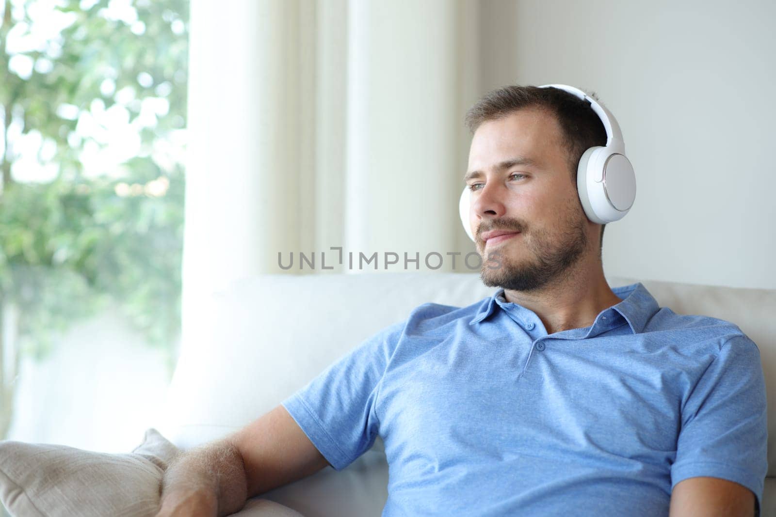 Satisfied man wearing headphone listening audio sitting on a sofa at home looking through a window