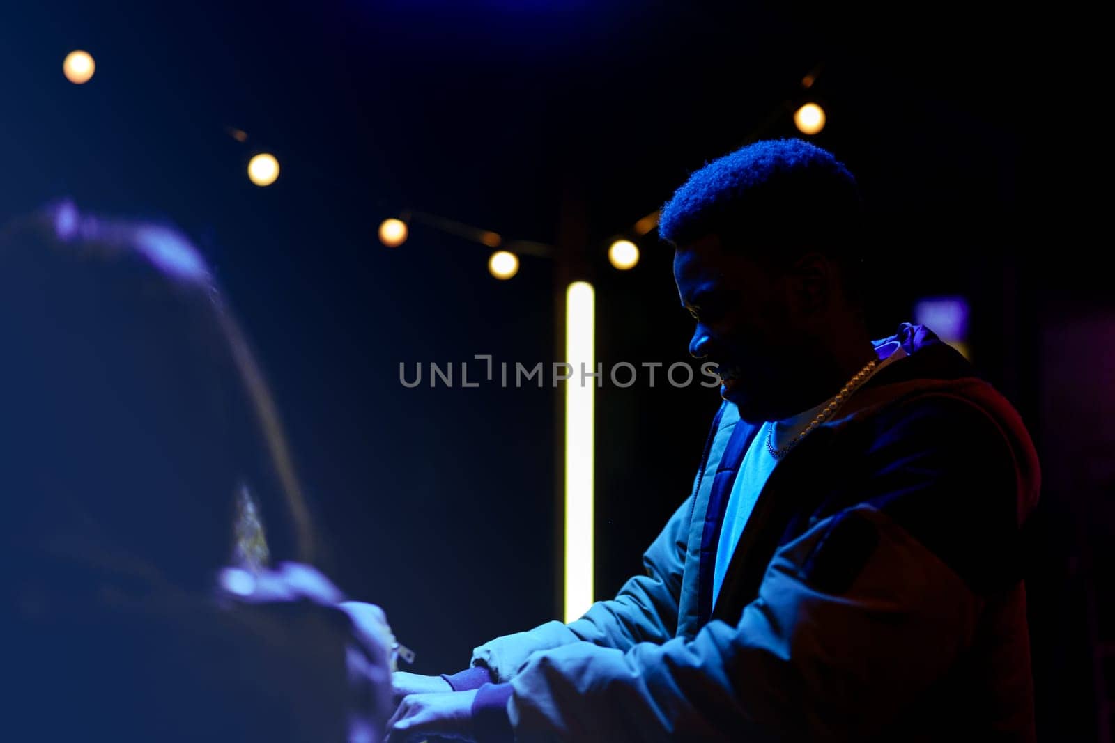 Two individuals interact in a dimly lit setting adorned with string lights. Their expressions reflect warmth and connection during a lively evening gathering.