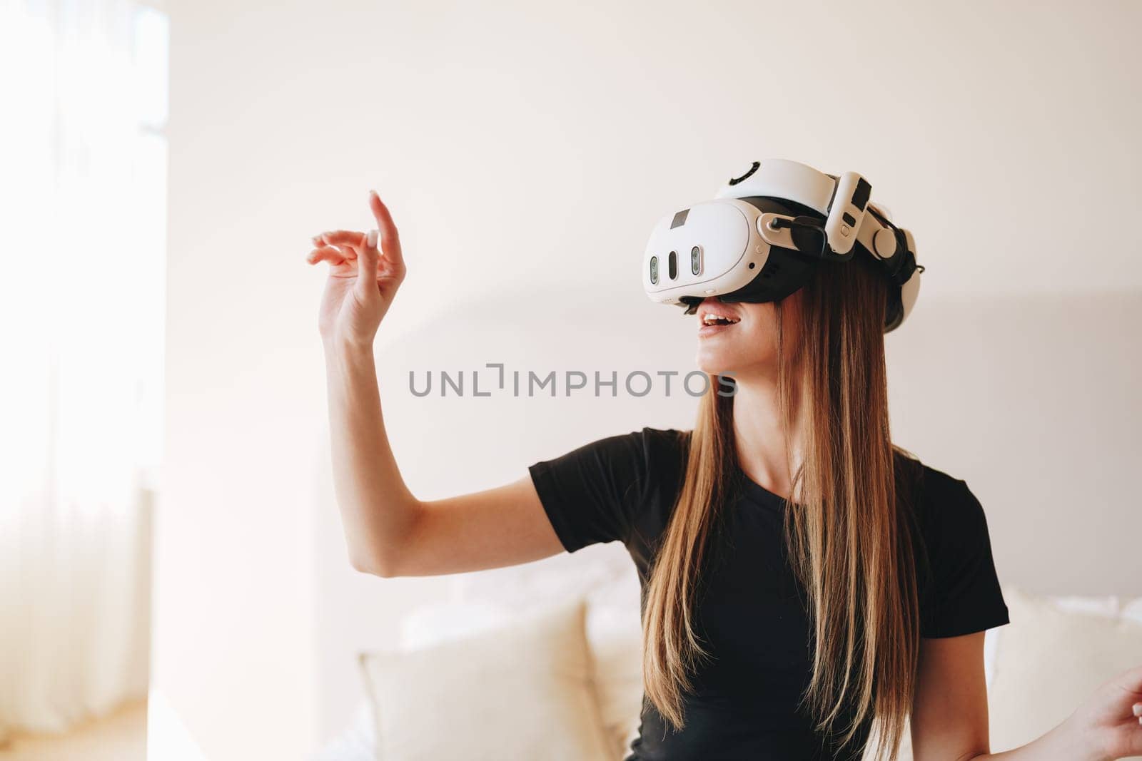 Engaged in a virtual reality experience, a young woman with long hair wears a headset and moves her hand as if reaching out to touch unseen objects while smiling in a well-lit room.