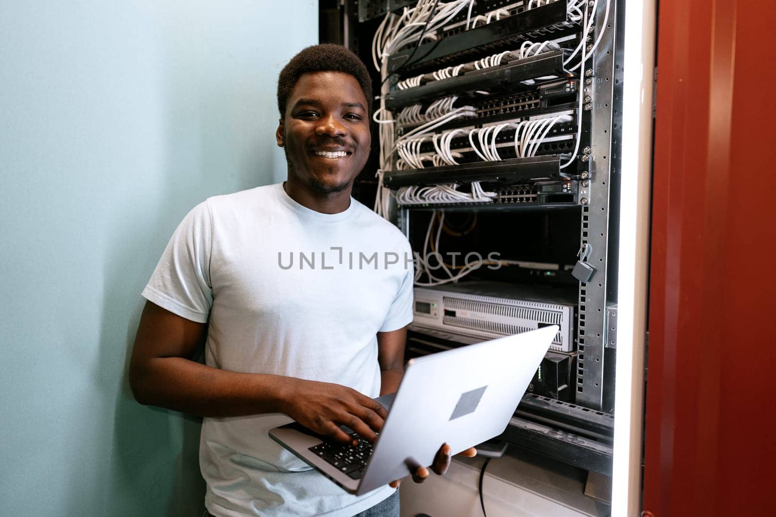 A young professional stands beside server racks, smiling widely while using a laptop. The environment is a data center, highlighted by multiple cables and equipment.