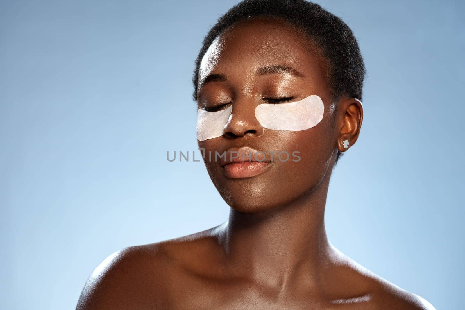 A woman is sitting peacefully with her eyes closed, applying under-eye patches to revitalize her skin. The serene atmosphere enhances her skincare routine in a studio setting.