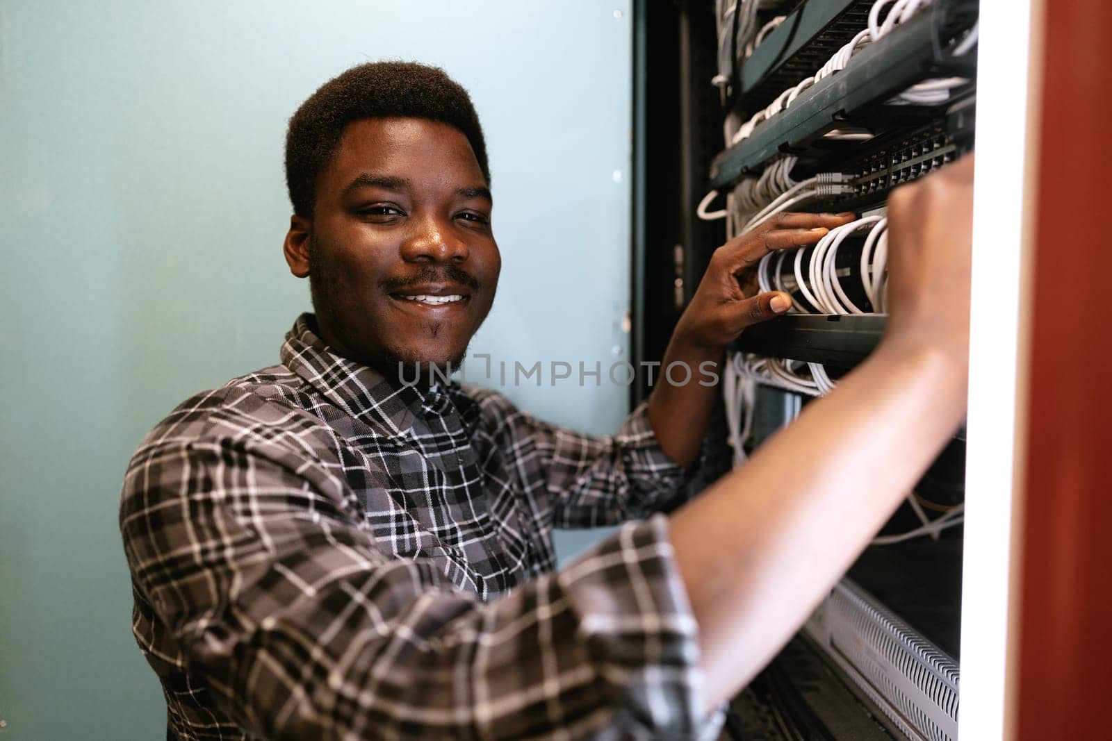 A technician smiles while managing network cables in a server room. The organized setup highlights his attention to detail in maintaining network infrastructure.