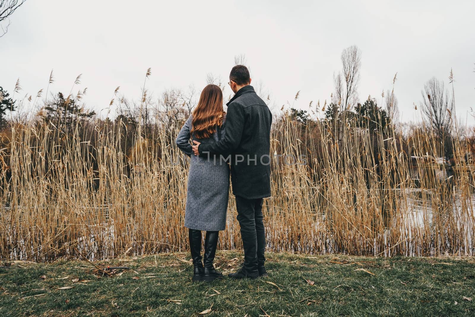 A couple stands close together, gazing at the water while surrounded by tall grass. The day is cloudy, creating a serene atmosphere for their intimate moment.