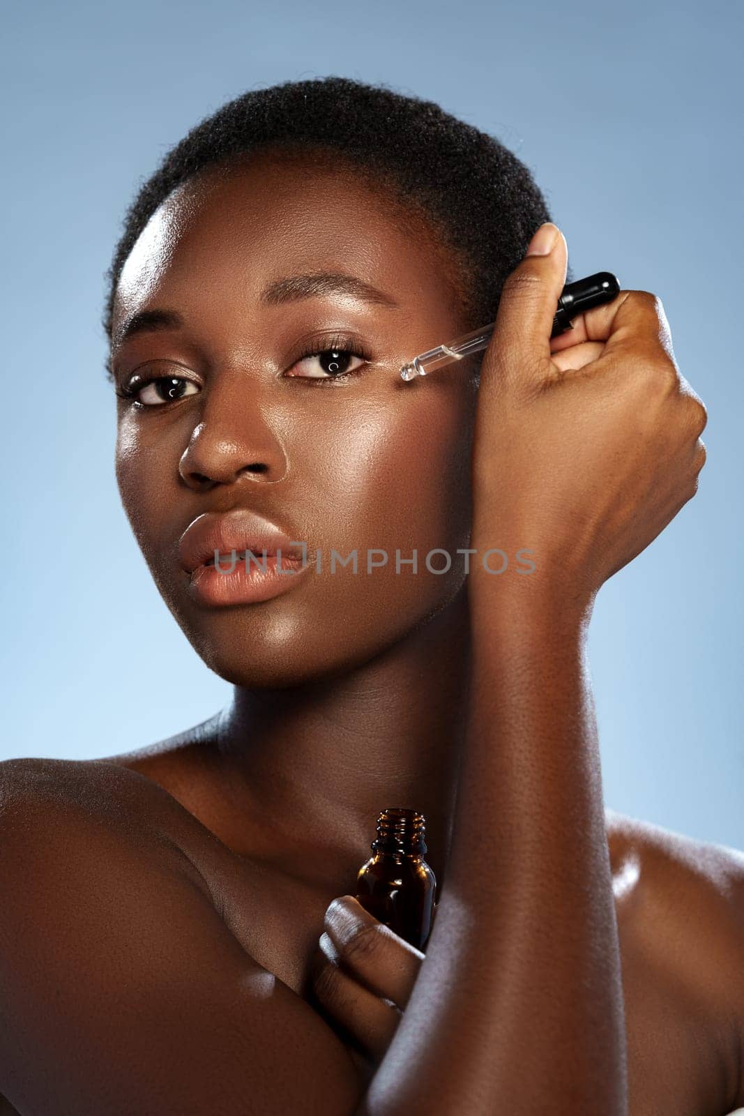 A woman with short, natural hair is delicately applying a serum to her cheek using a dropper. She showcases a glowing complexion in a well-lit studio with a blue background.
