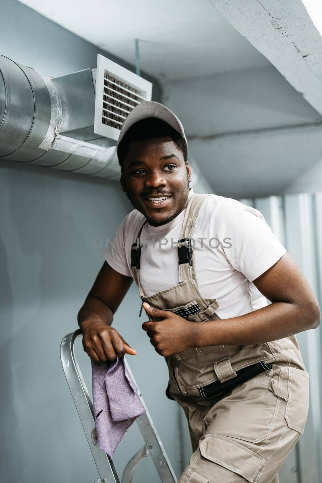 A young worker is on a ladder, smiling and holding a purple cloth. He is dressed in beige overalls and a cap, ready to perform maintenance in a well-lit indoor area.