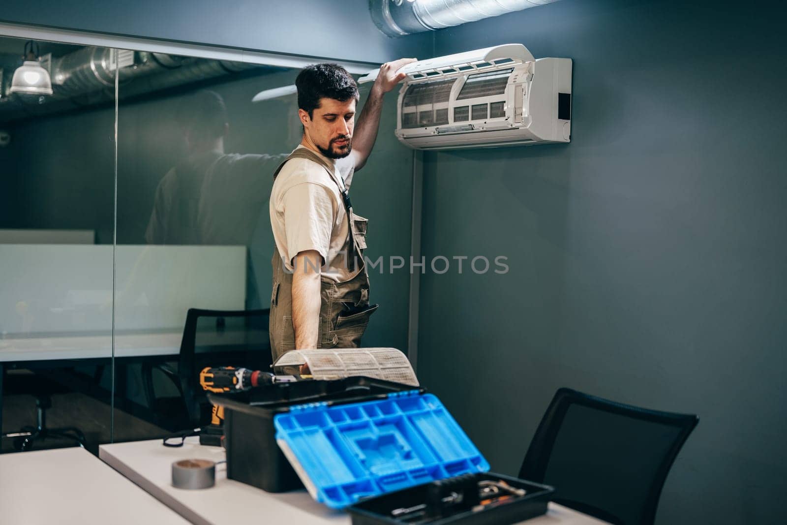 A technician is skillfully installing an air conditioning unit on the wall of a contemporary office. Tools and equipment are organized on the table nearby, showcasing a professional workspace.