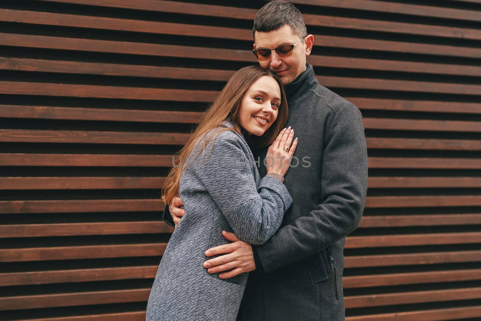 A couple enjoys a moment together, embracing warmly while standing in front of a wooden slatted wall.