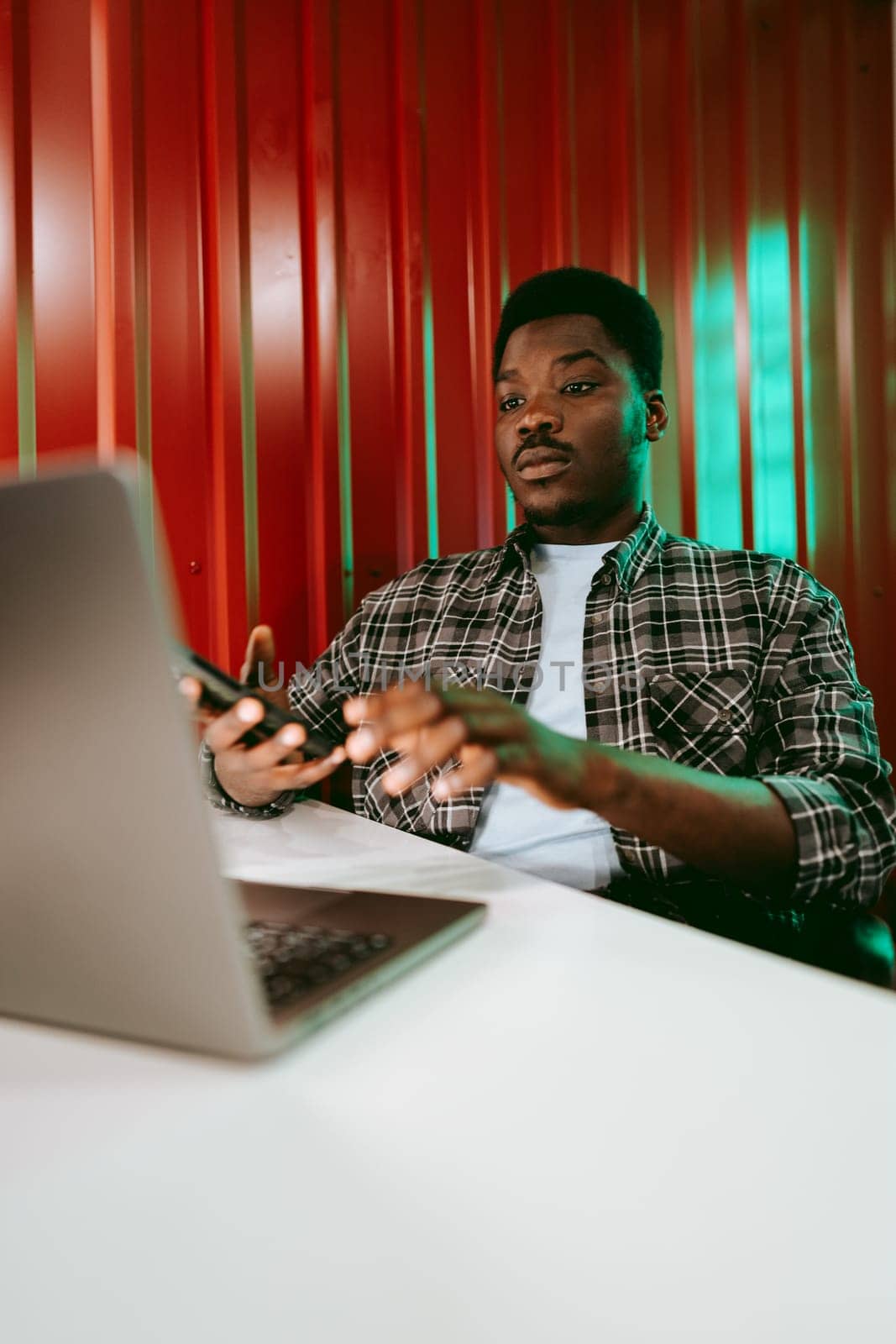 A young man sits at a table, intently typing on a laptop and checking his smartphone in a colorful office environment with red backdrop.