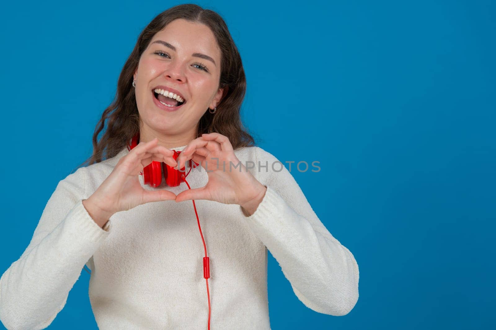 Young woman in white sweater and red headphones smiling while making a heart shape with her hands against a blue background, representing kindness, connection, love, and positive emotions. Copy space