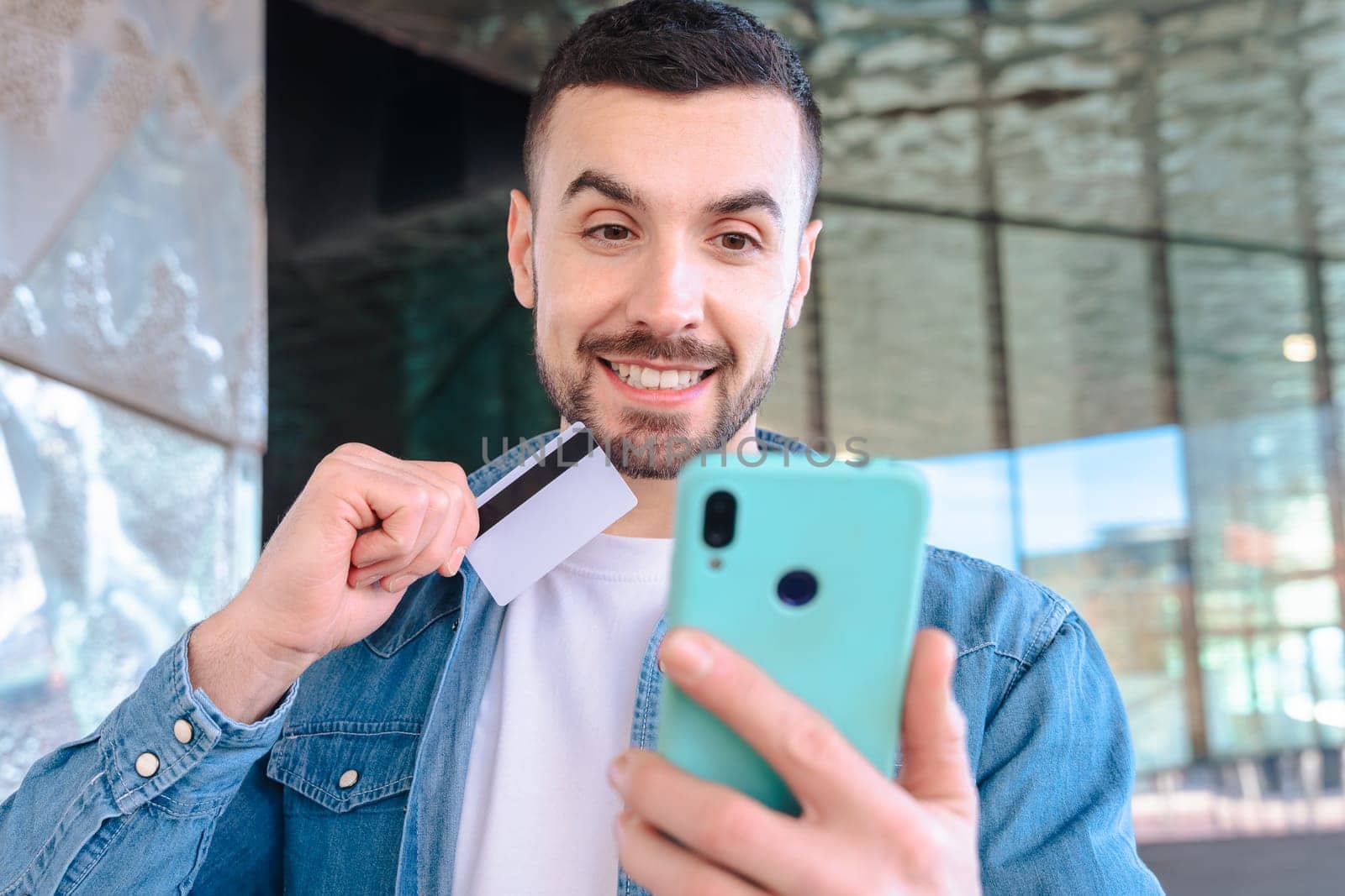 Happy young man using his smartphone while holding a credit card. He smiles with excitement, standing outside a glass building, enjoying an online shopping or payment experience.