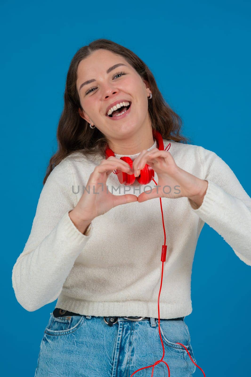 Happy young woman in white sweater and red headphones smiling while making a heart shape with her hands against a blue background, representing kindness, connection, love, and positive emotions. Vertical