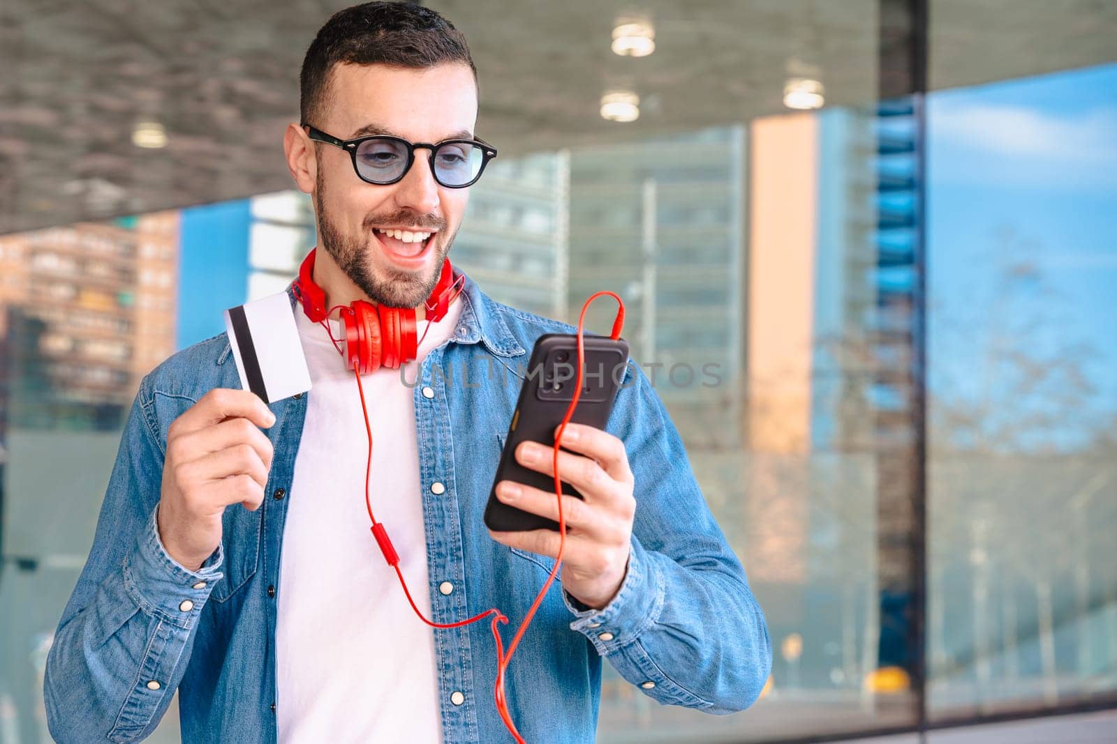 Happy man celebrating a successful online purchase while holding a credit card and smartphone. He smiles with excitement, wearing headphones and sunglasses in a modern urban environment.