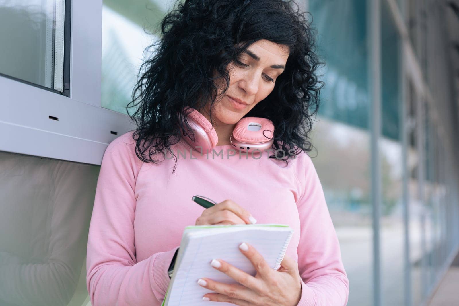 Caucasian sporty woman in a pink hoodie writing in a notebook outdoors. Wearing headphones around her neck, she appears focused and relaxed after a workout in an urban setting.