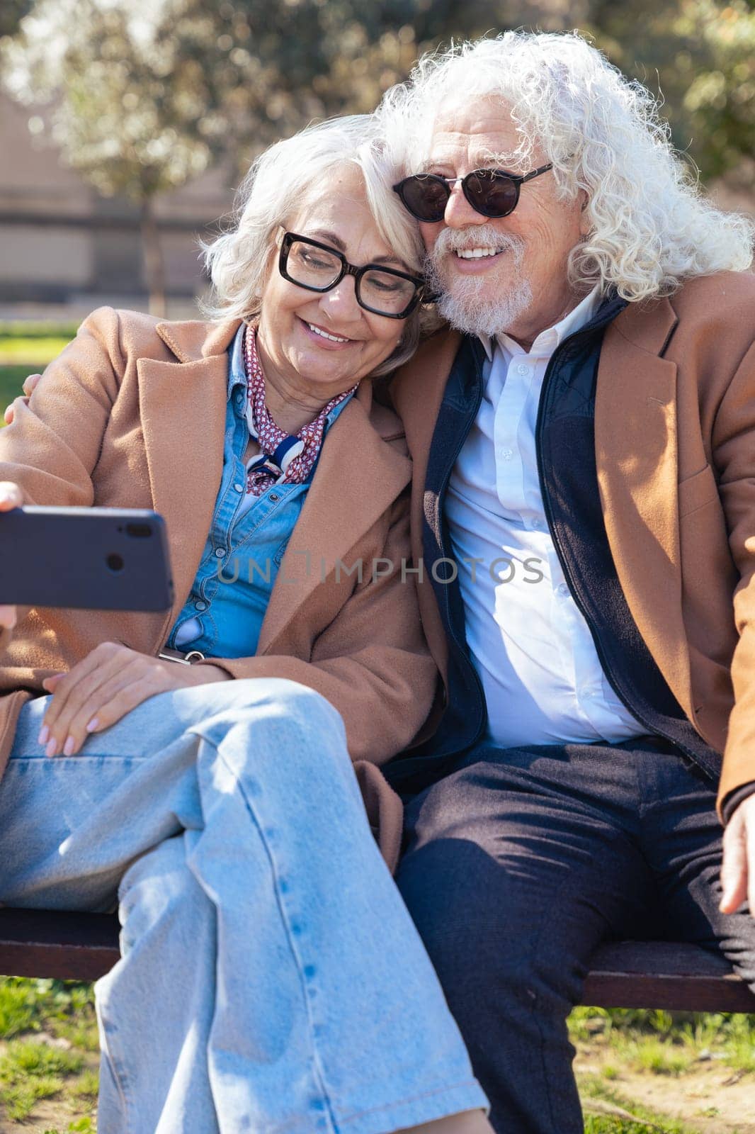 Elderly couple dressed in brown coats taking a cheerful selfie outdoors. They smile while holding a smartphone, capturing a happy moment together in a sunny and casual environment.Vertical