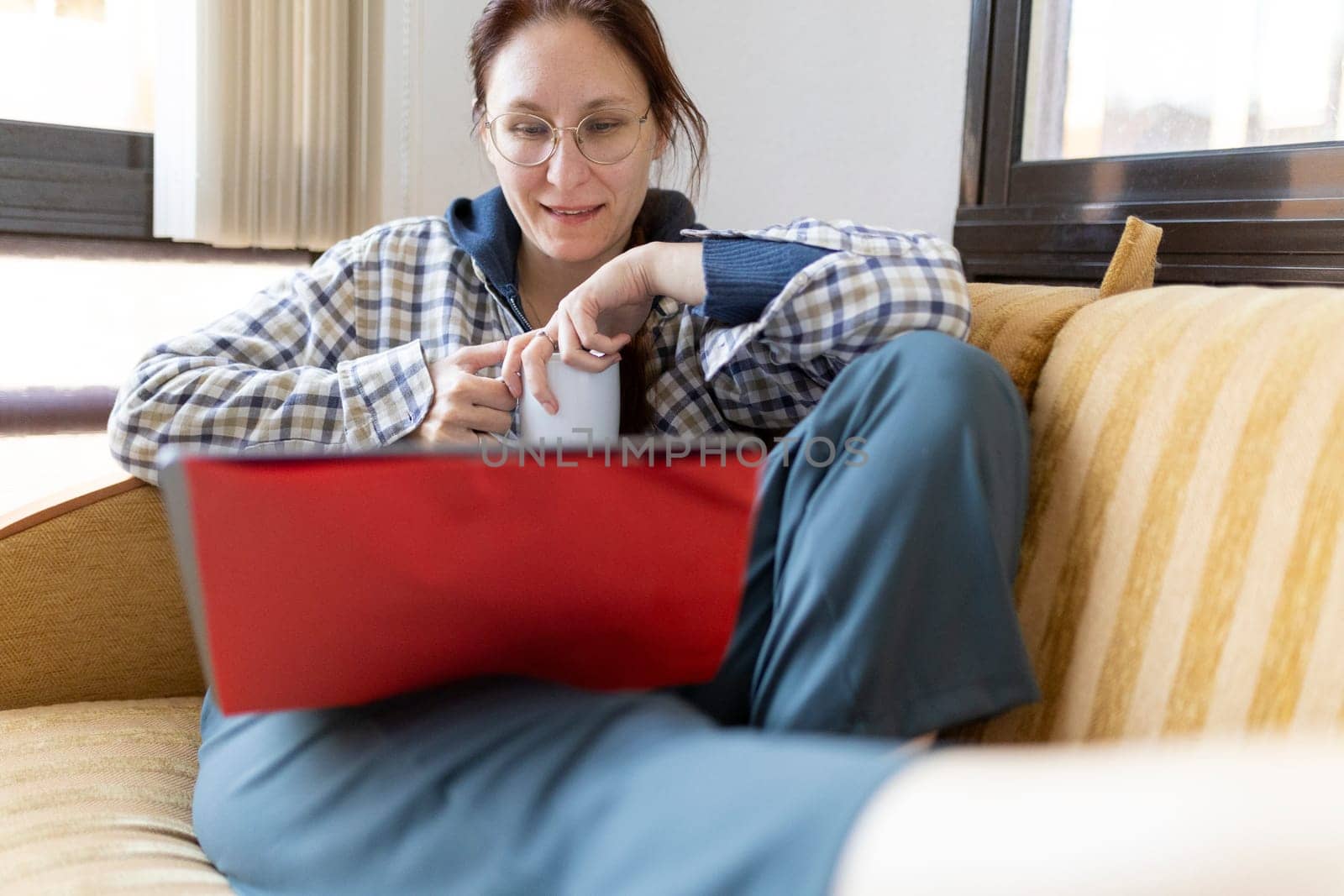 Woman relaxing on sofa using laptop and drinking tea, working from home
