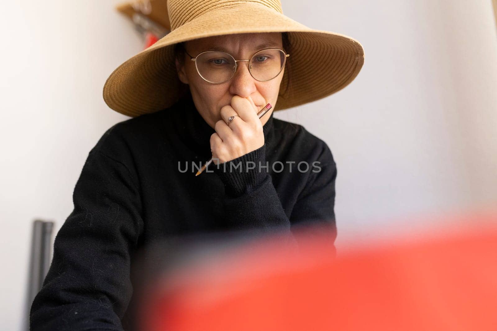 Woman artist wearing straw hat and eyeglasses concentrating while holding paintbrush in her home studio