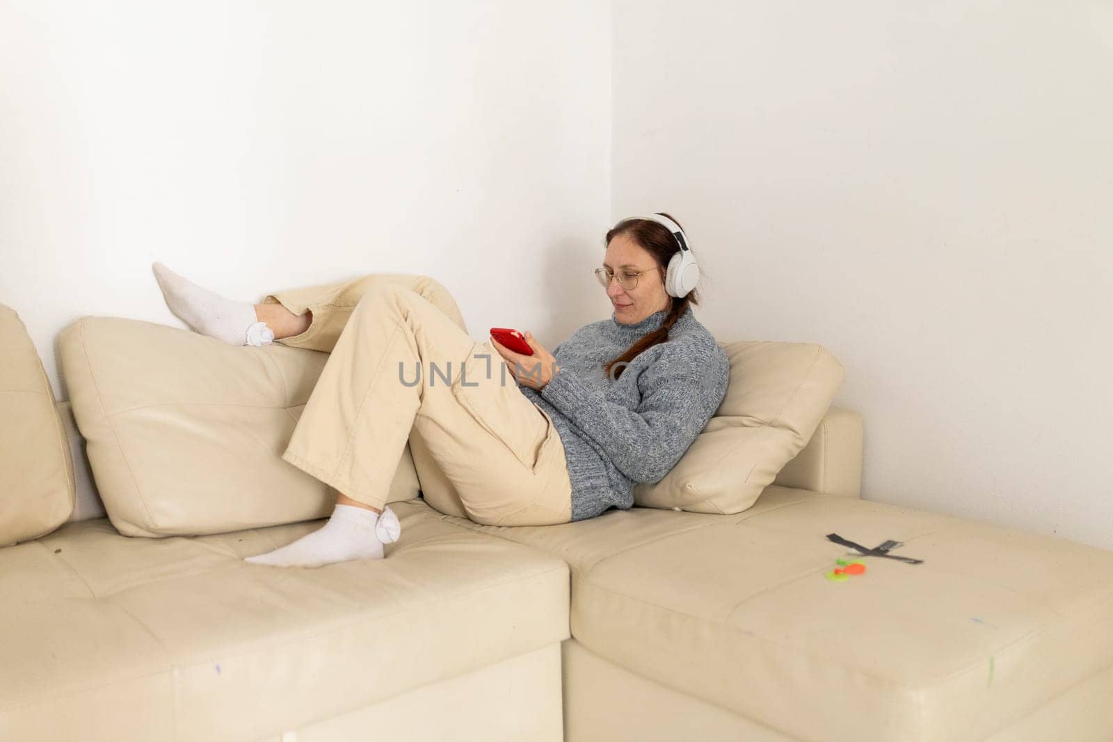 Woman wearing headphones is lying on a sofa, using smartphone and listening to music
