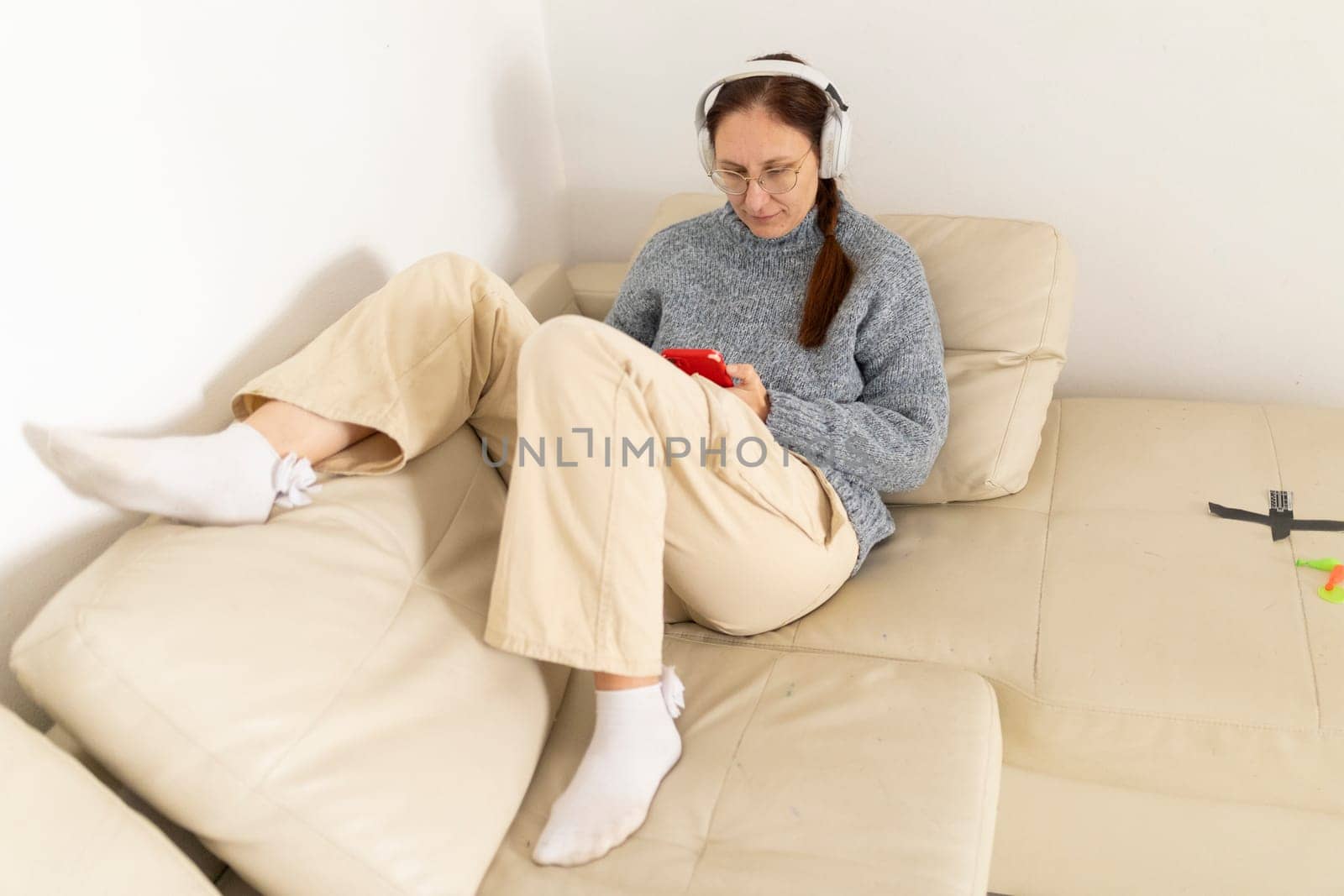 Woman wearing headphones enjoying music and using smartphone while relaxing on comfortable sofa at home