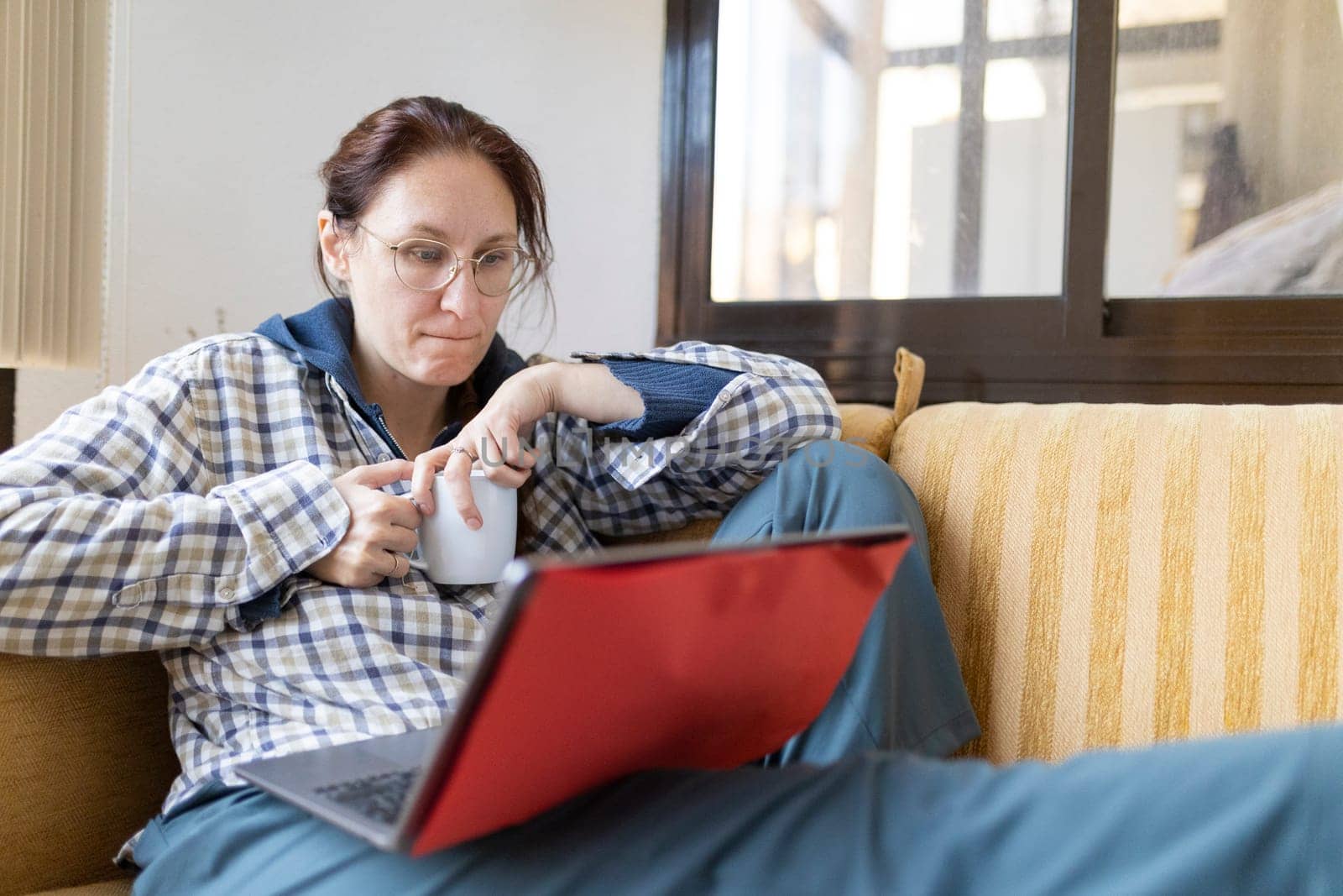 Woman sitting comfortably on sofa, enjoying coffee while working remotely using laptop