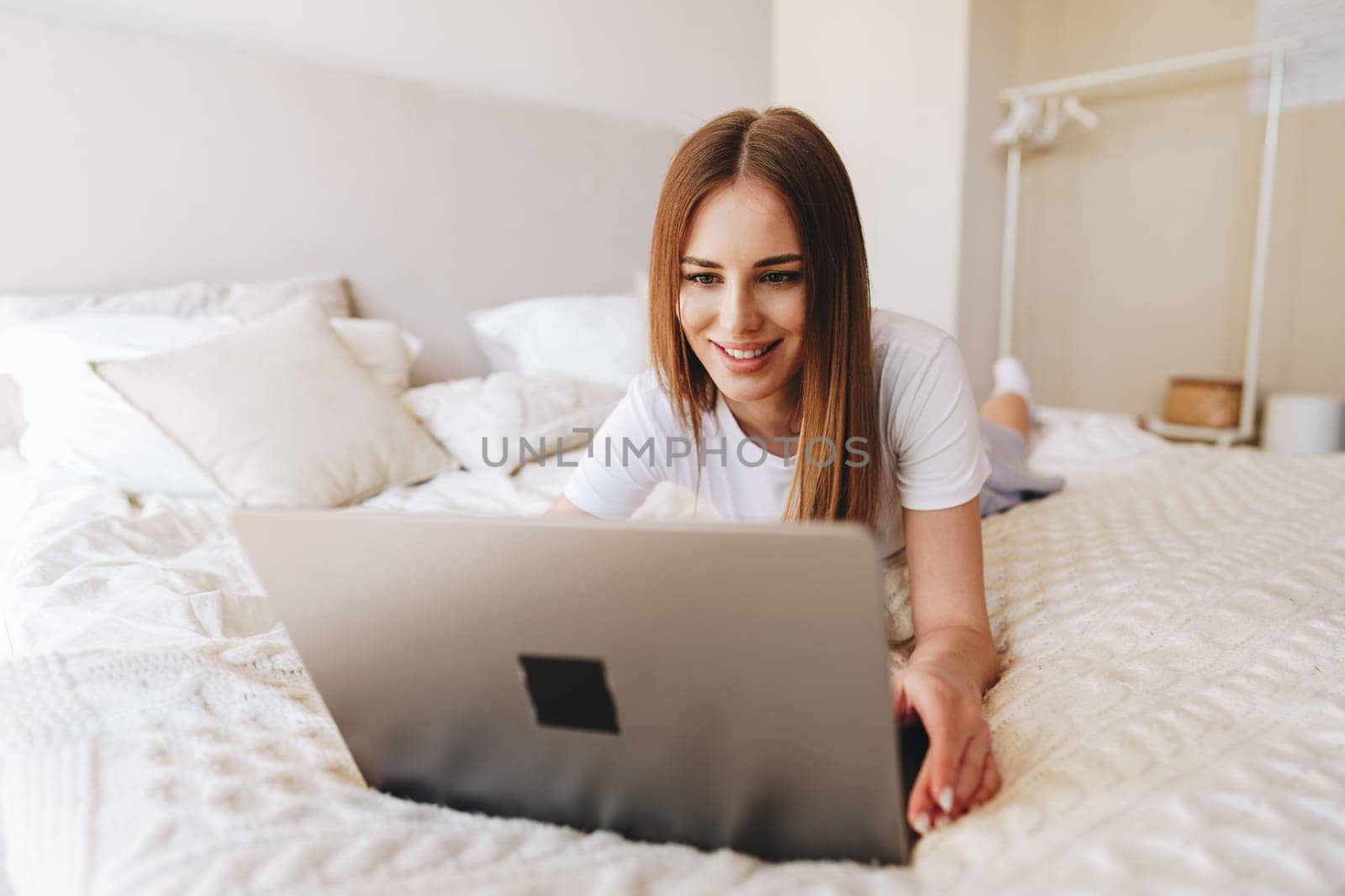 A woman with long brown hair smiles while using a laptop on her bed. Soft pillows and a warm ambiance create a comfortable and inviting atmosphere in her personal space.