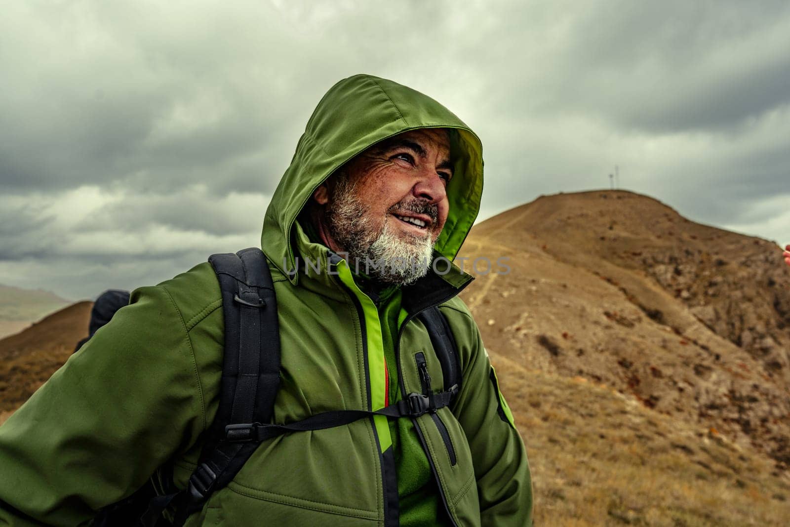 A determined man wearing a green jacket smiles as he navigates the hilly landscape under a moody sky, showcasing a spirit of adventure and exploration in nature.