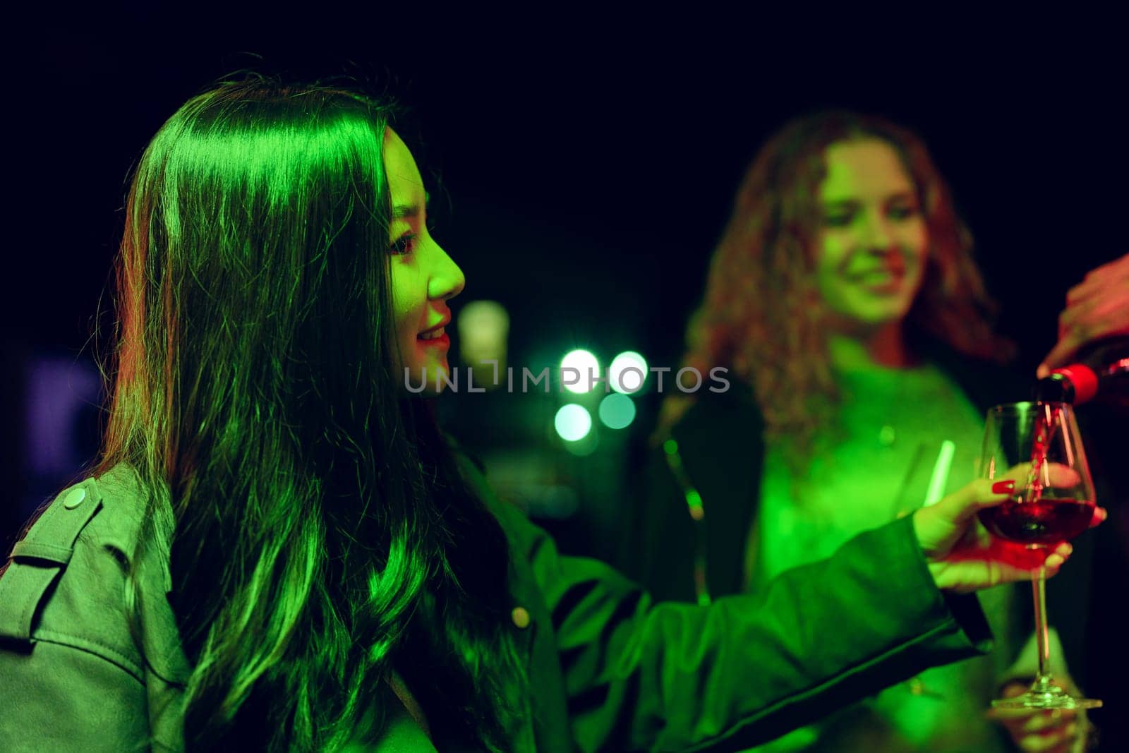 Two friends share a joyful moment during an energetic night gathering. One is pouring wine into a glass under colorful lights, highlighting their smiles and laughter.